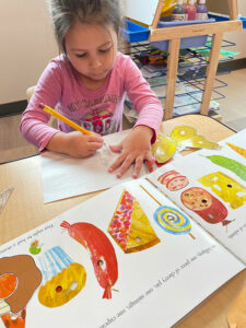 Girl writes on paper while looking The Very Hungry Caterpillar book 